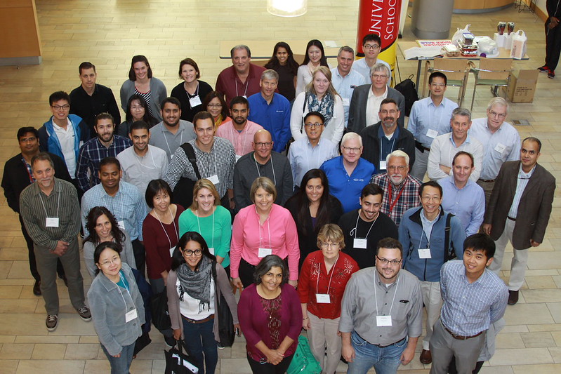 Large group of adults standing together indoors in several rows, looking up toward the camera. They are wearing conference name badges, and tables with refreshments are visible in the background